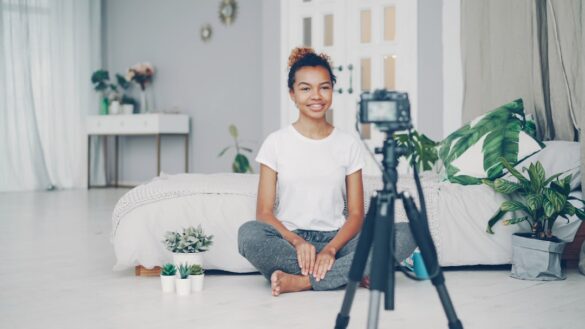 Good-looking African American girl creative blogger is recording video about plants sitting on floor of her modern apartment and talking looking at camera on tripod.