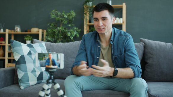 Happy guy vlogger is broadcasting video content using smartphone camera at home talking gesturing waving hand. Vlogging, people and lifestyle concept.